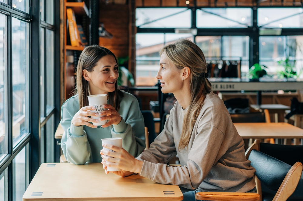 Two,Women,Sitting,In,A,Coffee,House,Talking,And,Drinking